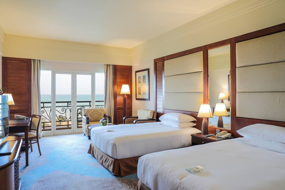 A hotel room with two beds, a desk, a chair, and a balcony with a view.
