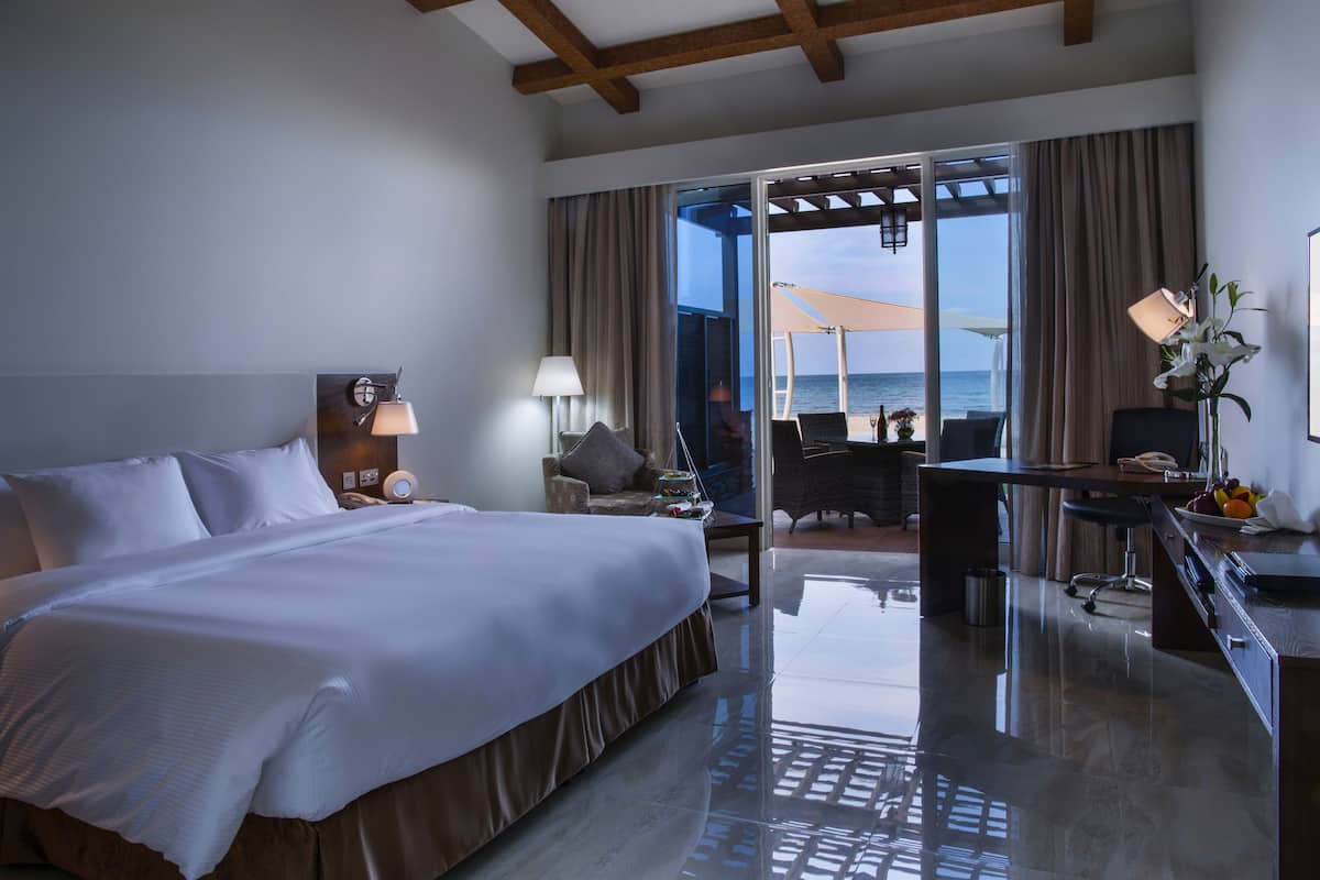 A spacious hotel room with a large bed, a desk, and a view of the sea through a sliding glass door.