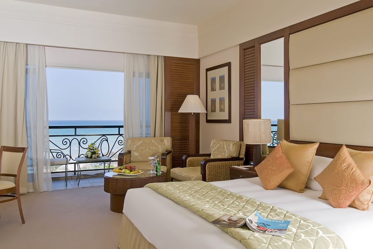 A hotel room with a bed, two armchairs, a small table with a fruit plate, a lamp, and a view of the ocean.