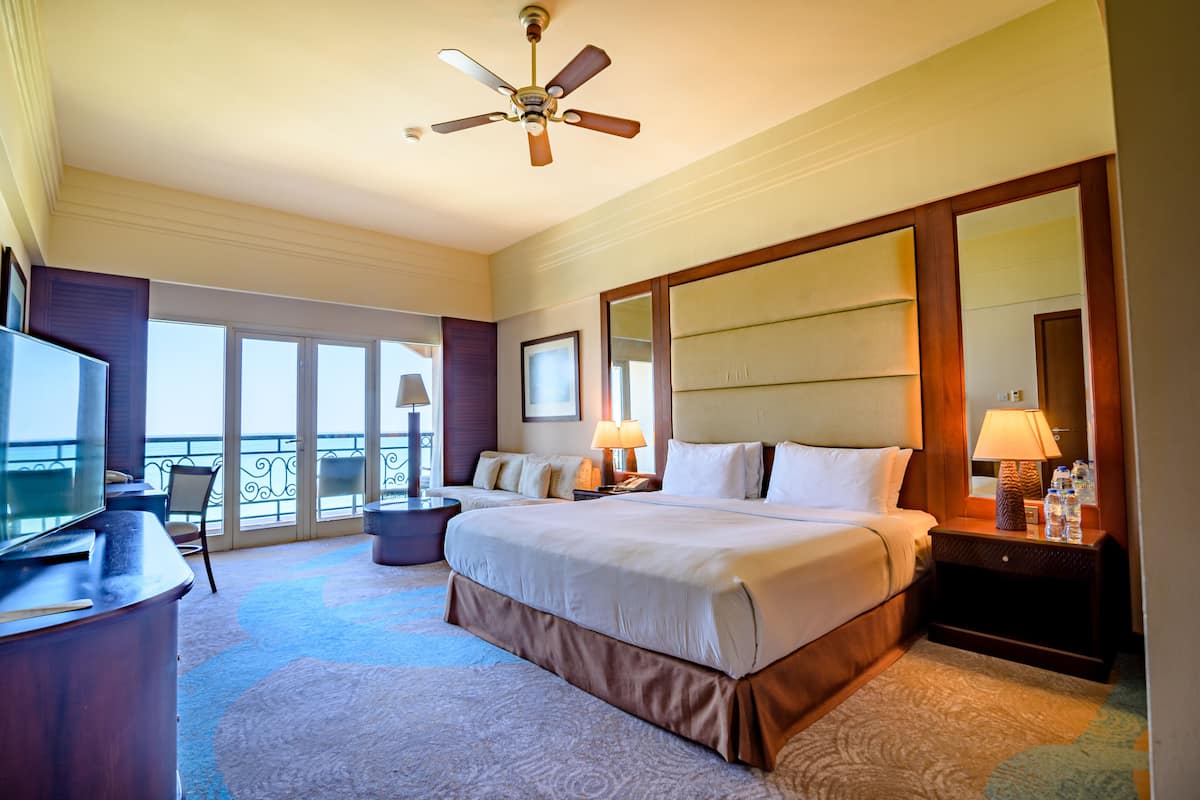 A hotel room with a large bed, a desk with a television, a balcony with a view, and a ceiling fan.