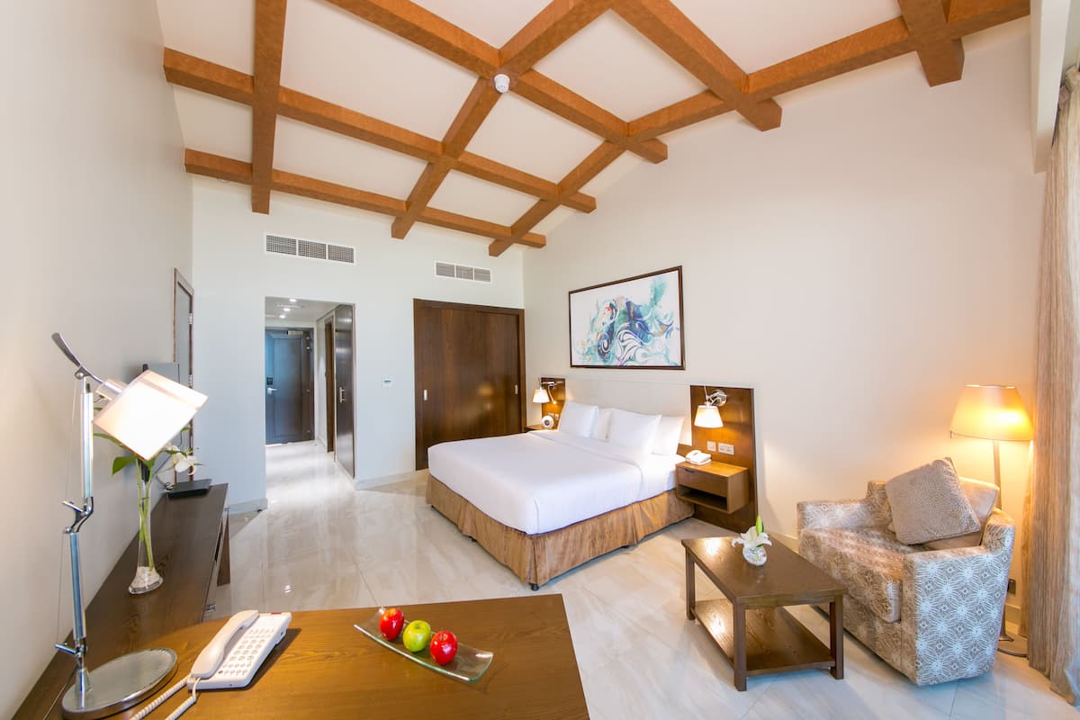 A hotel room with a large bed, bedside tables, a desk with a telephone and fruit, a sofa, and a painting on the wall.