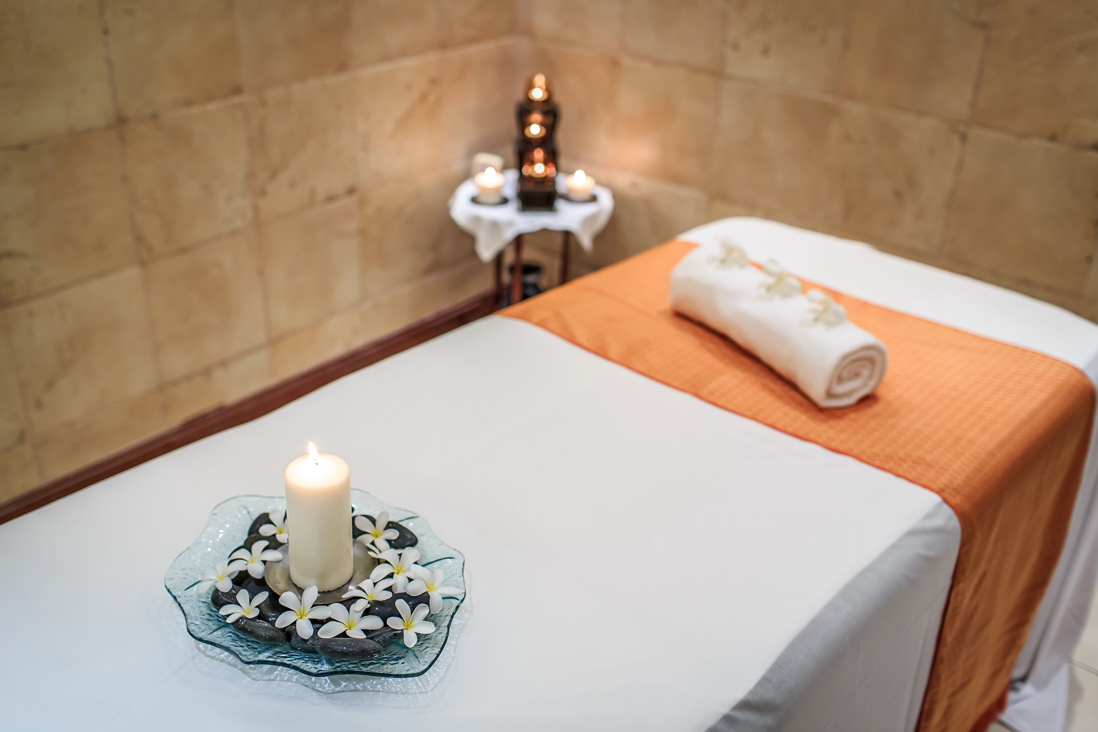 A spa room with a white bed, an orange towel, a lit candle, and a small table with candles in the background.