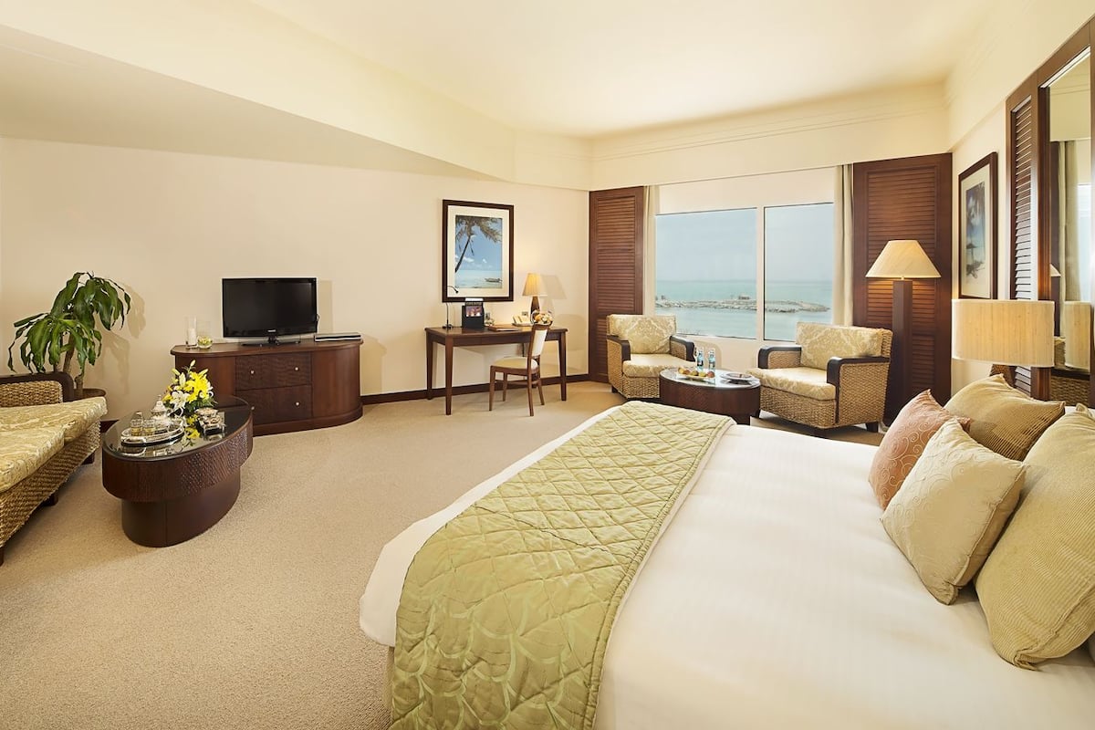 A hotel room with a large bed, a sofa, a TV, a desk, and a view of the sea.