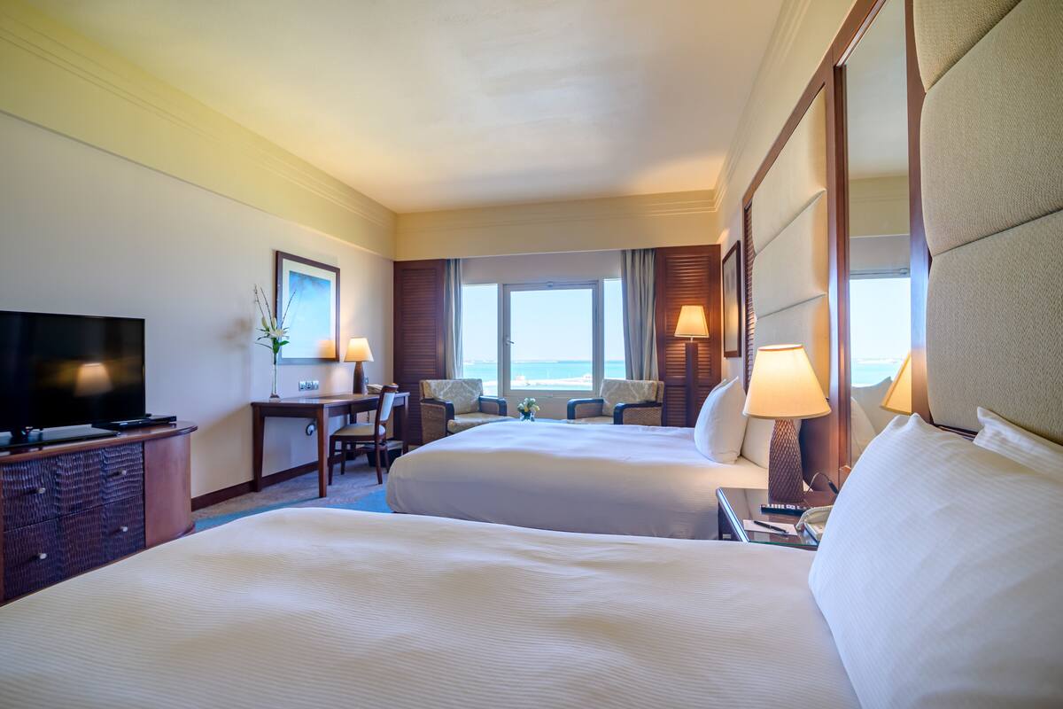 A hotel room with two beds, a desk, a TV, and a view of the ocean.