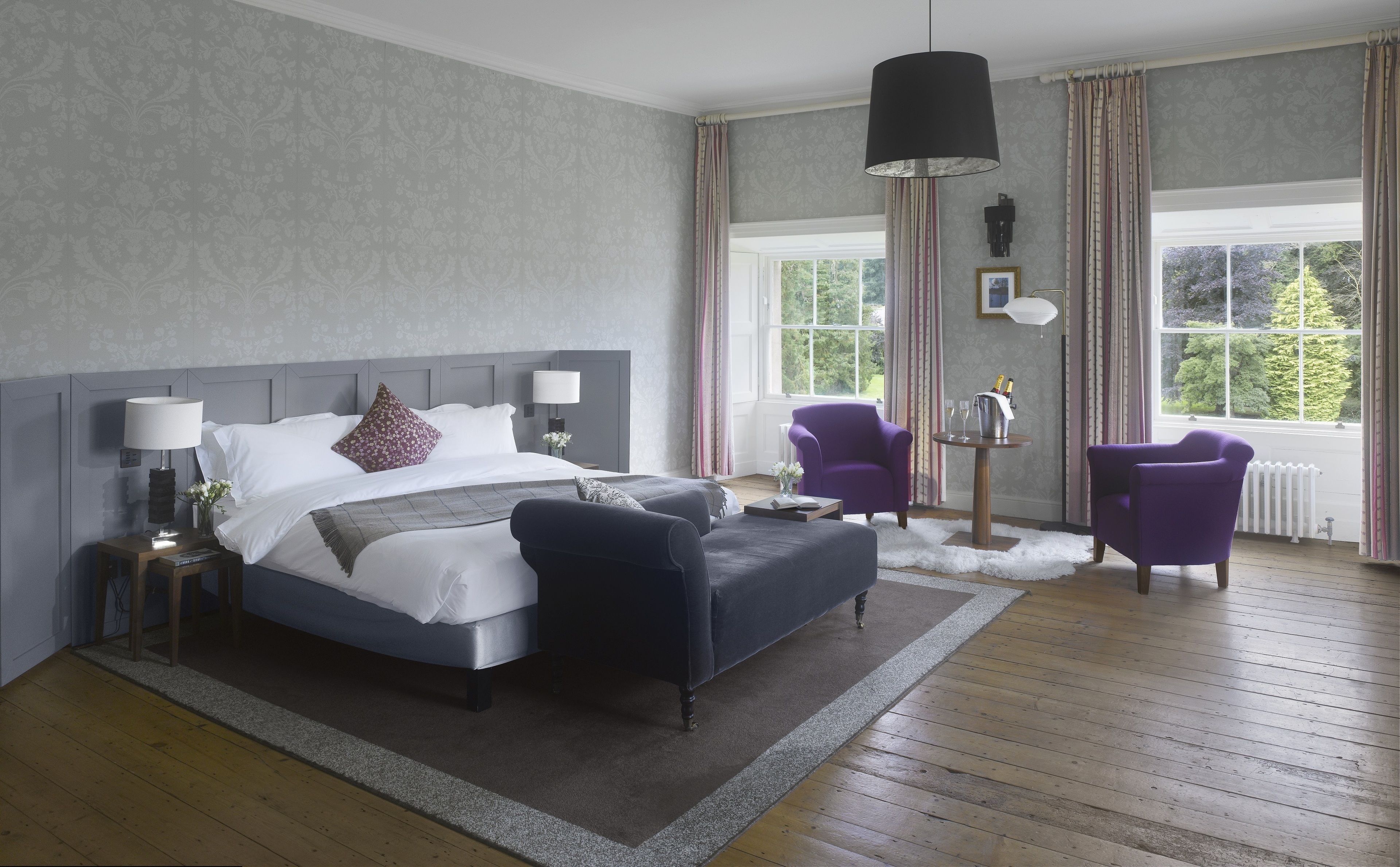 A modern bedroom with a large bed, two chairs, a small table, and a window with curtains.