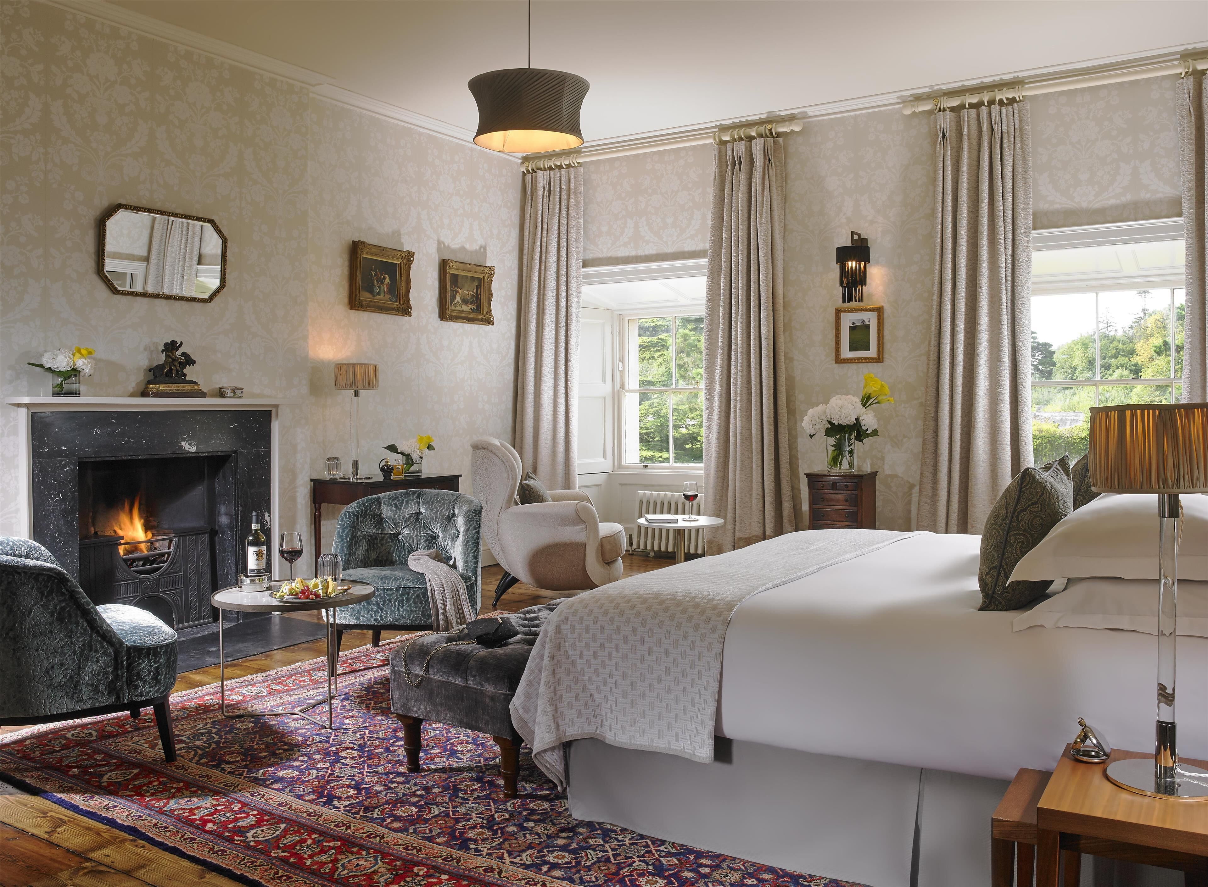 Une chambre d’hôtel avec une cheminée, un grand lit, un coin salon avec des fauteuils, une table à manger et une vue sur la verdure environnante.