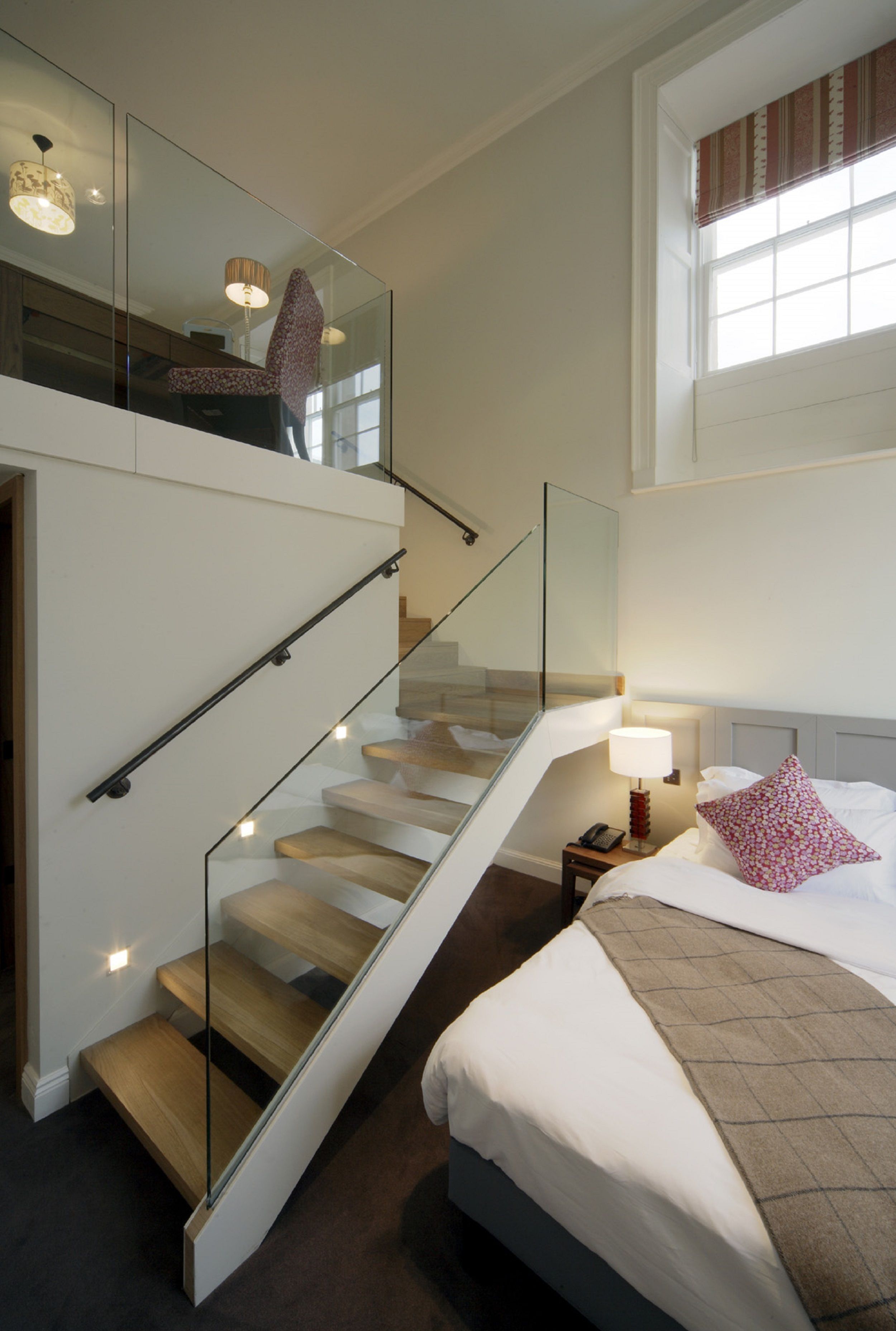 Una habitación de hotel moderna con una escalera de cristal, una cama grande, una mesita de noche con lámpara y una ventana con cortinas.