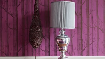 Ein Zimmer mit violettem Wandteppich, einem hängenden Schmuckstück, einer Lampe und einem Bett mit violetter Decke und Kopfkissen.