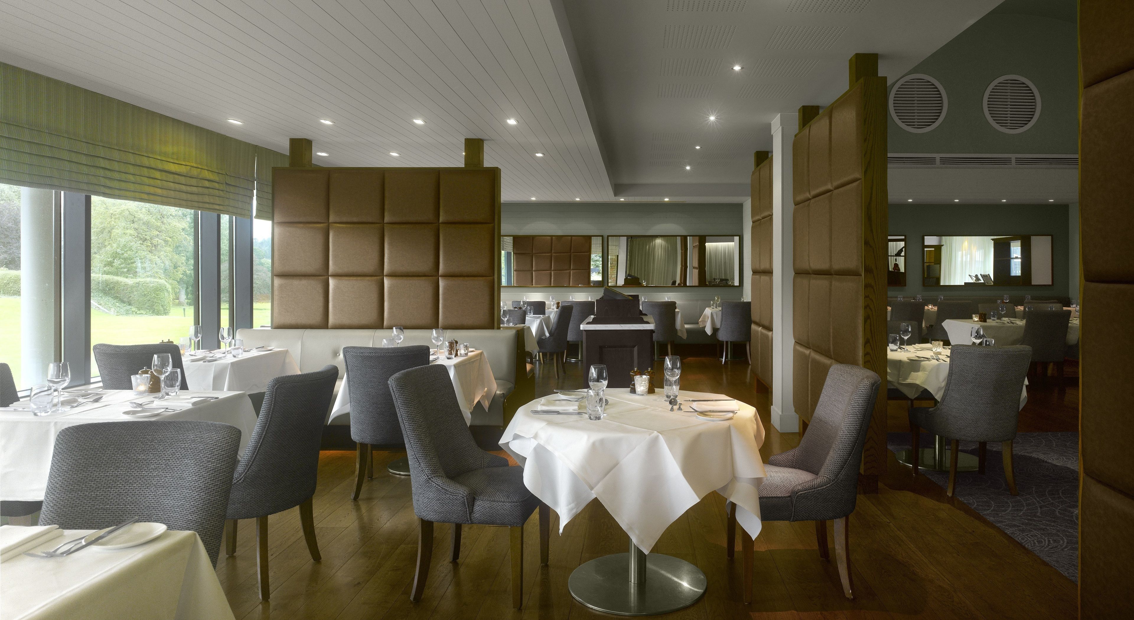 Un restaurante con varias mesas y sillas, un techo alto con iluminación empotrada y grandes ventanales que ofrecen vistas al exterior.