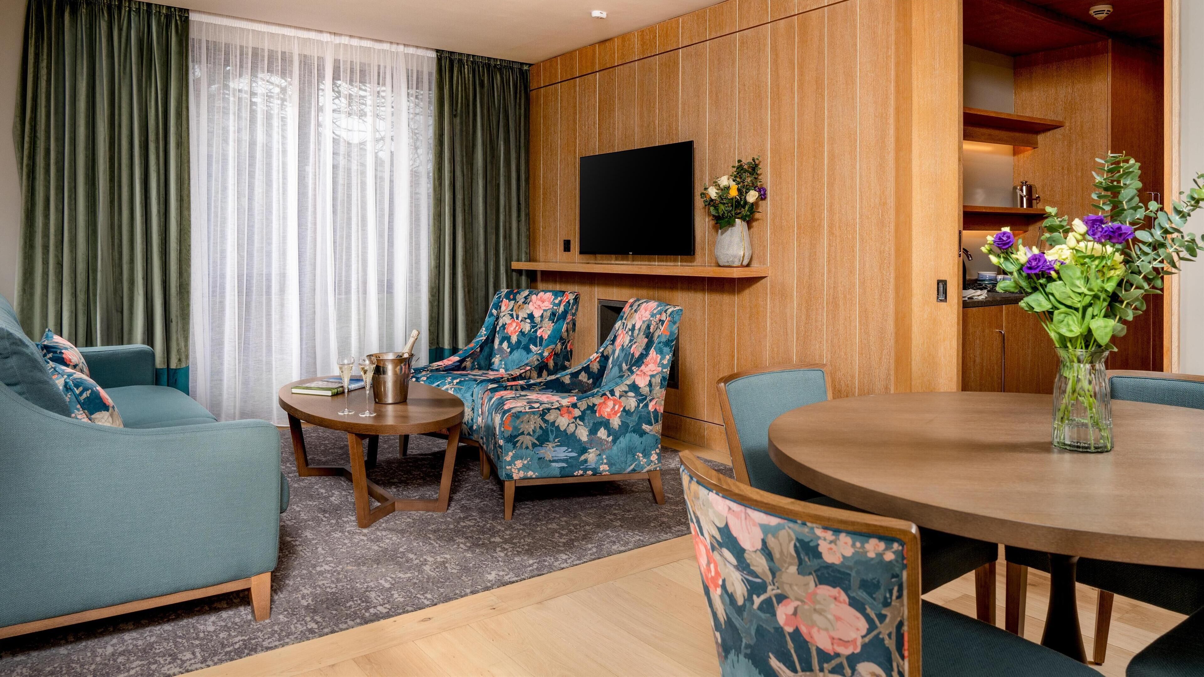 A modern living room with a sofa, two floral armchairs, a coffee table, and a dining table with a vase of flowers.