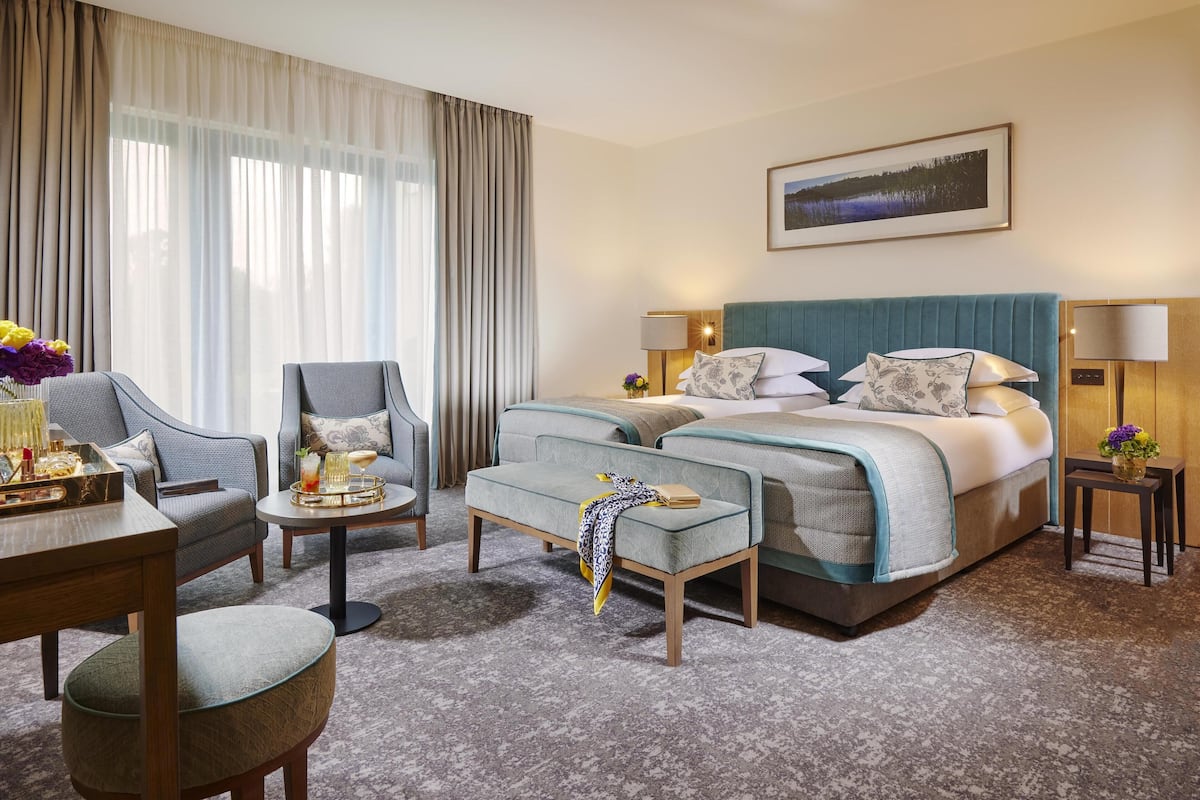 A hotel room with a large bed, two armchairs, a small table with a tray, and a view of a lake through the window.