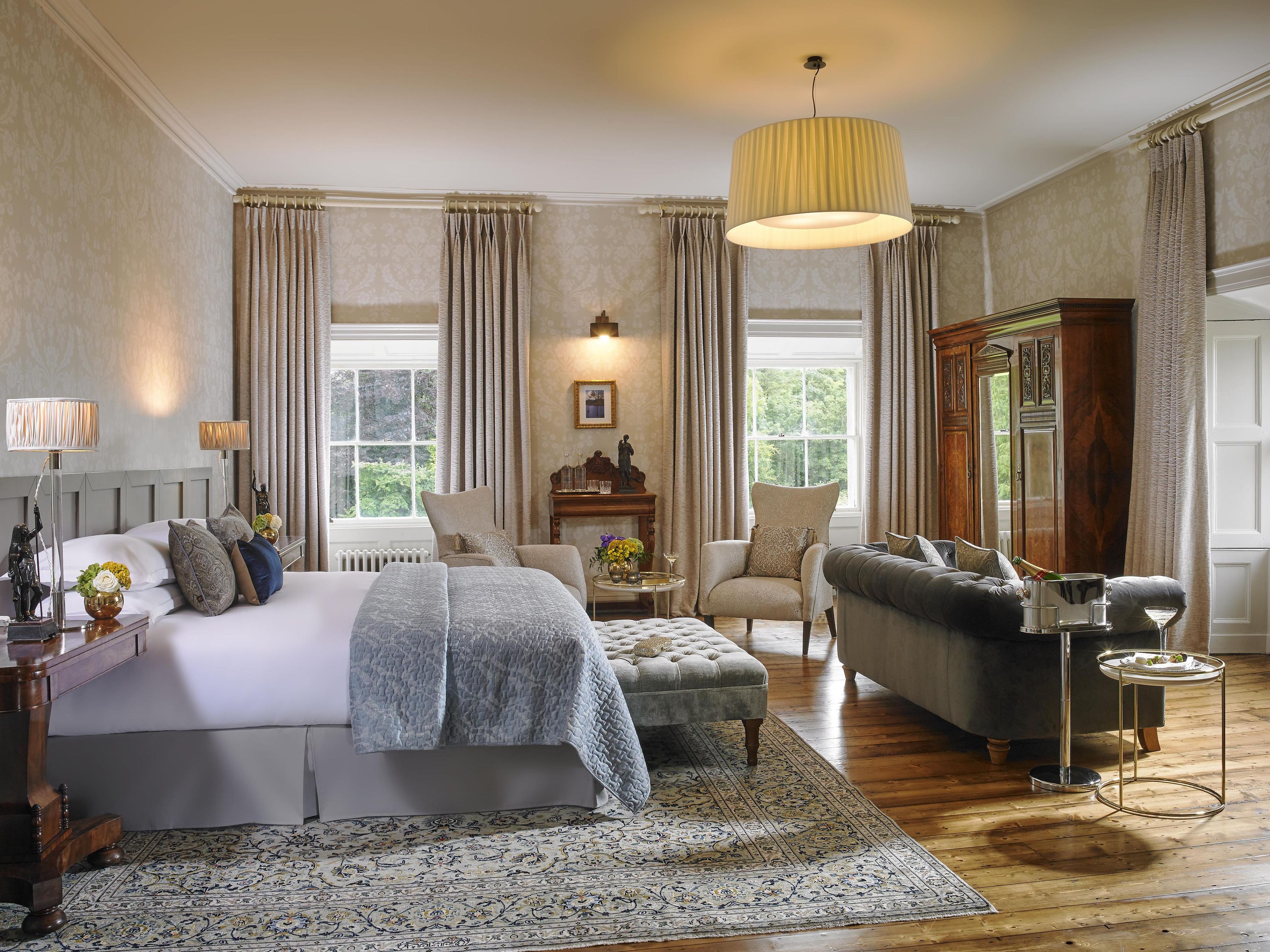 Um quarto espaçoso com uma cama grande, um espaço de estar com sofá e poltrona, piso de madeira e uma janela ampla que oferece iluminação natural.