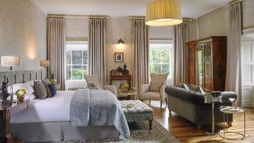 Um quarto espaçoso com uma cama grande, um espaço de estar com sofá e poltrona, piso de madeira e uma janela ampla que oferece iluminação natural.
