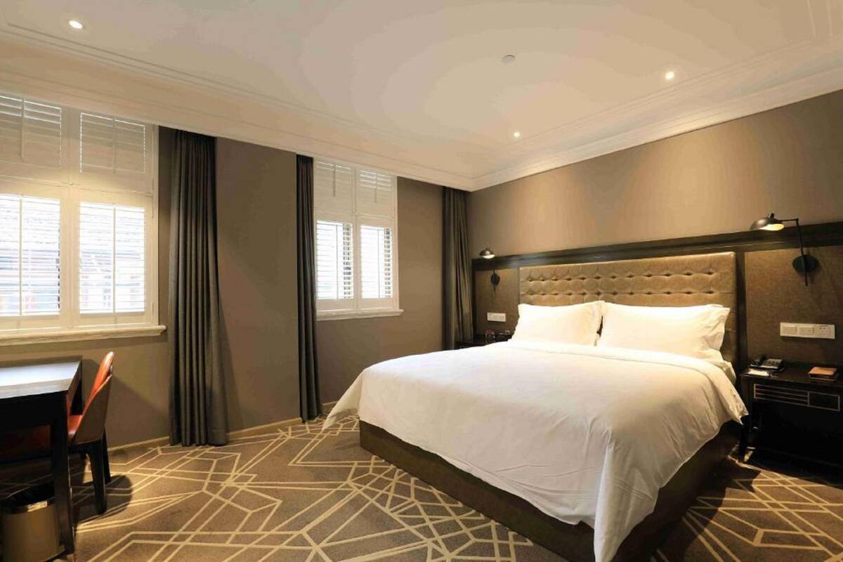Habitación de hotel con una cama grande, un escritorio y una silla. Ventanas con cortinas, cabecero y una lámpara fijada a la pared.