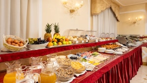 A buffet with a variety of food items and beverages, including fruits, vegetables, and prepared dishes, arranged on a long table with a red tablecloth. - Hotel A LA Commedia (Venice)