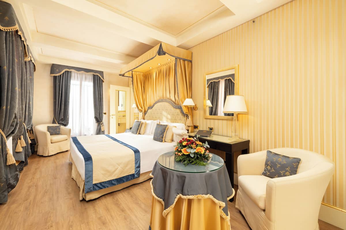 A hotel room with a large bed, a sitting area with a chair, a round table with a flower arrangement, and a desk with a lamp.