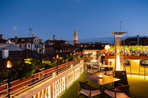 An outdoor terrace with tables and chairs, a railing, and a view of a historic building with a clock tower. - Hotel A LA Commedia (Venice)