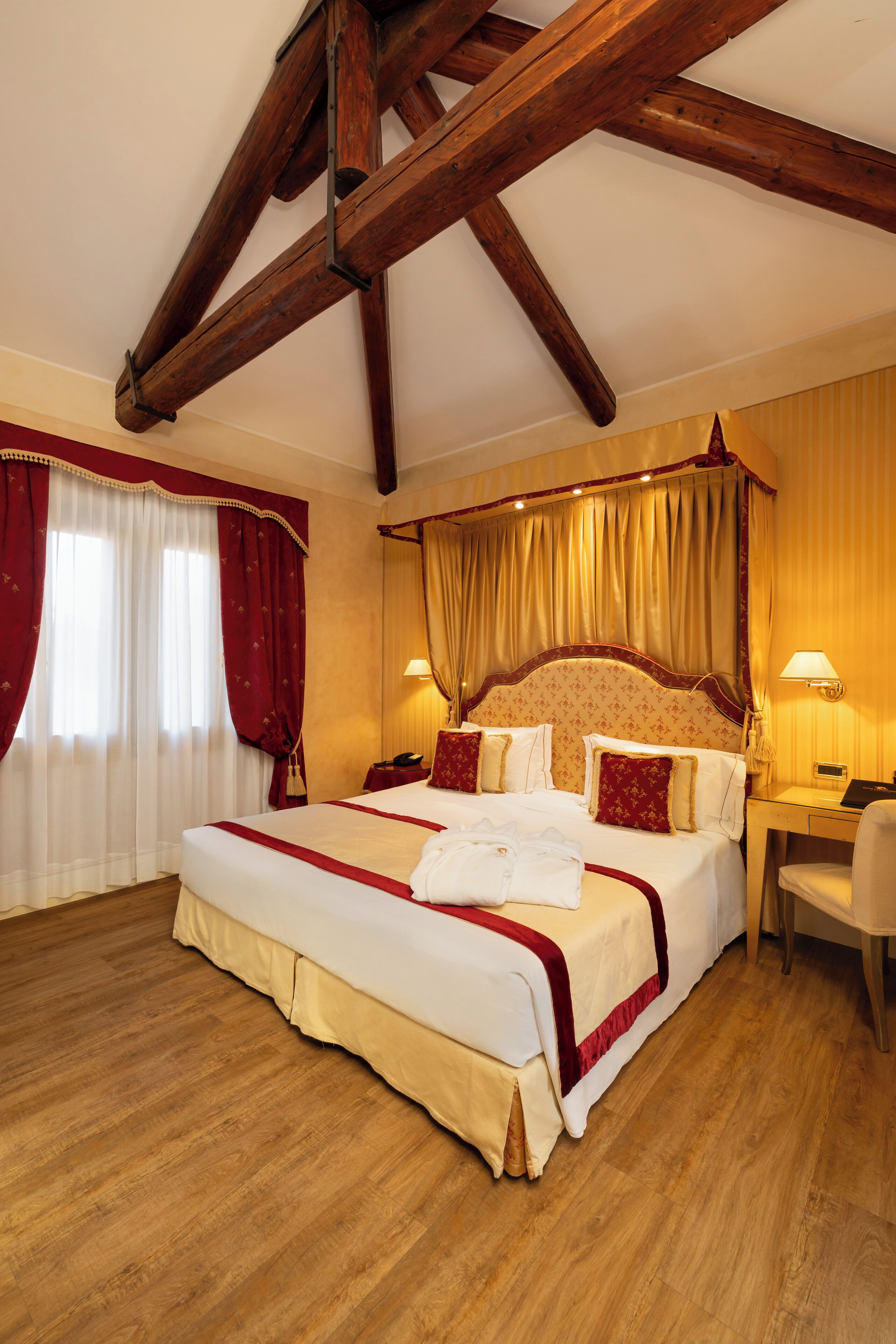 Un letto grande con baldacchino, travi di legno a vista al soffitto, una scrivania con una sedia e una finestra con tende.