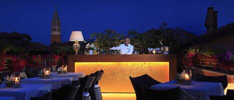 Zona de bar con un bartender, mesas con velas y una vista de una torre al fondo.
