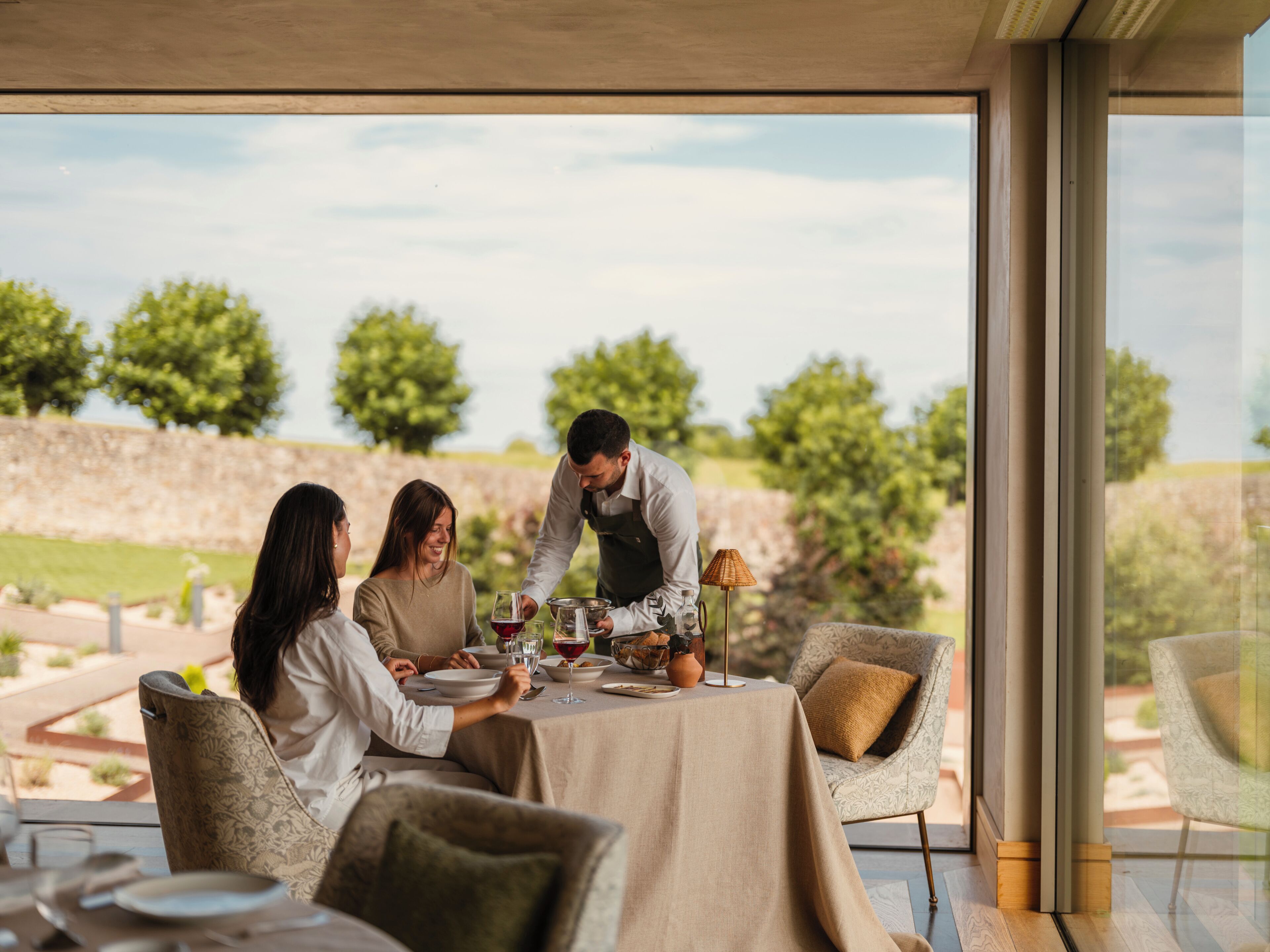 Un comedor con una mesa lista para tres personas, un camarero atendiendo y una vista de un viñedo al fondo.