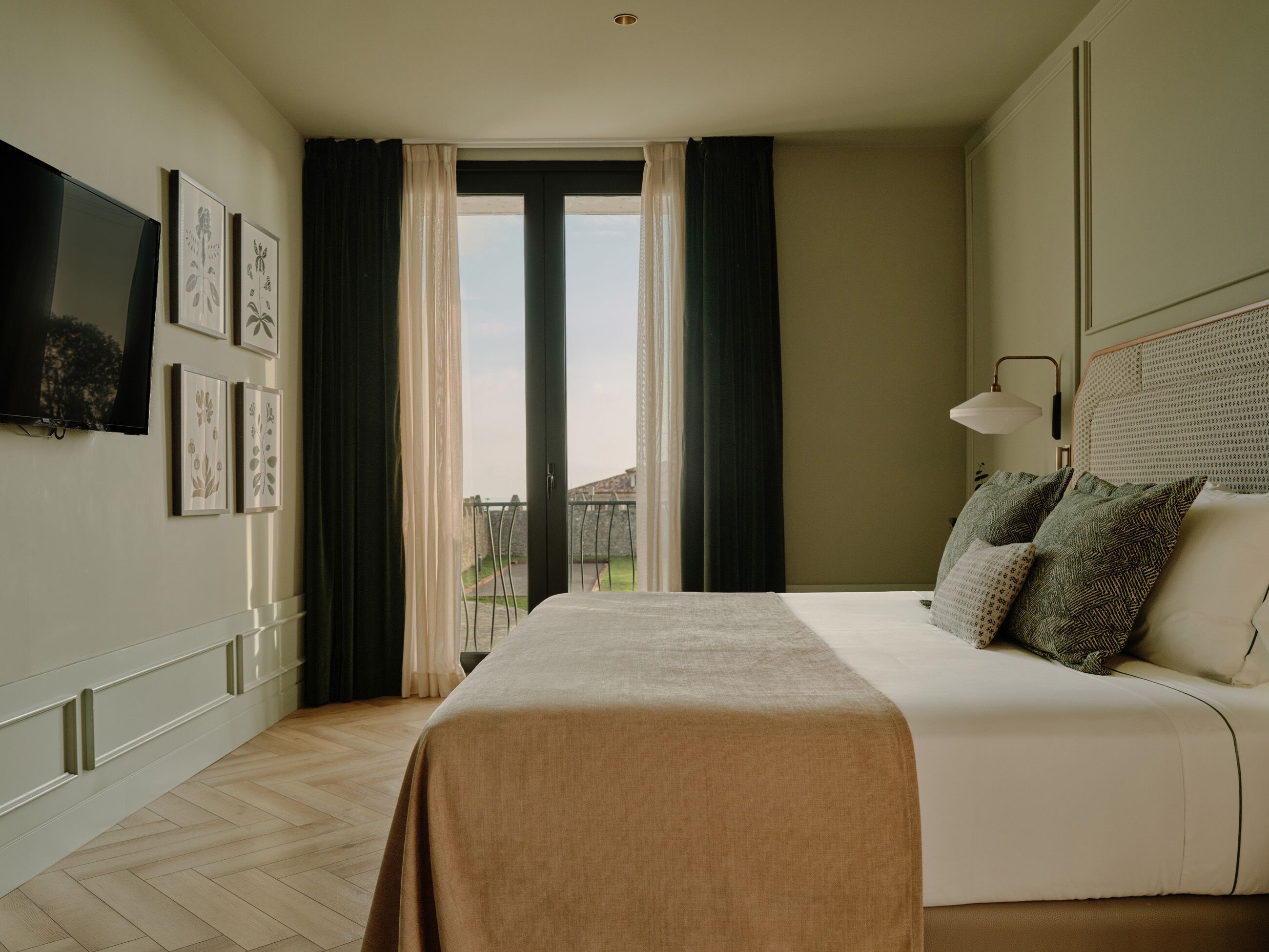A hotel room with a bed, a TV, a window with curtains, and artwork on the wall.