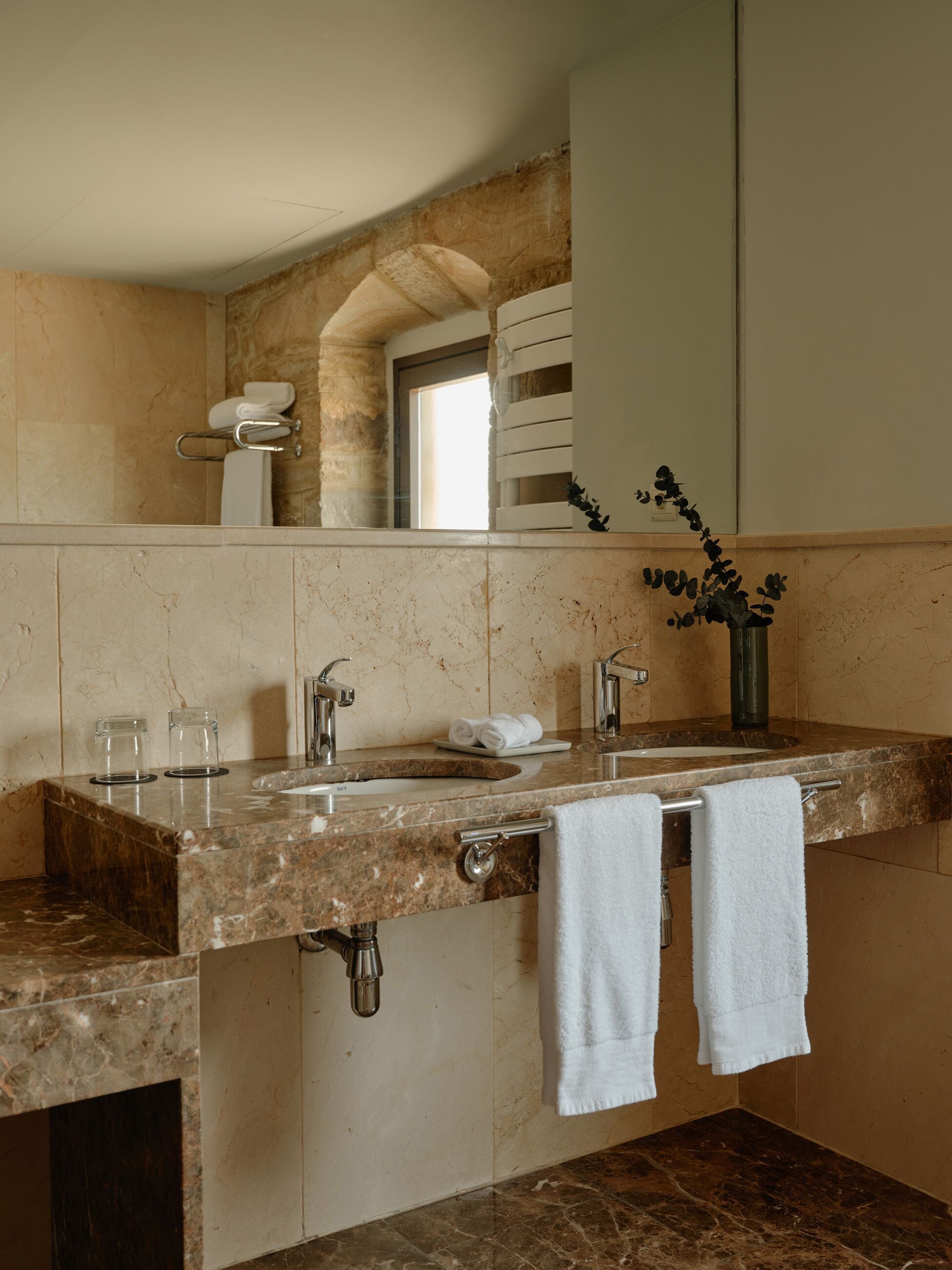 Un baño con lavamanos de mármol, dos toalleros, un tarro de cristal y un florero con una planta.