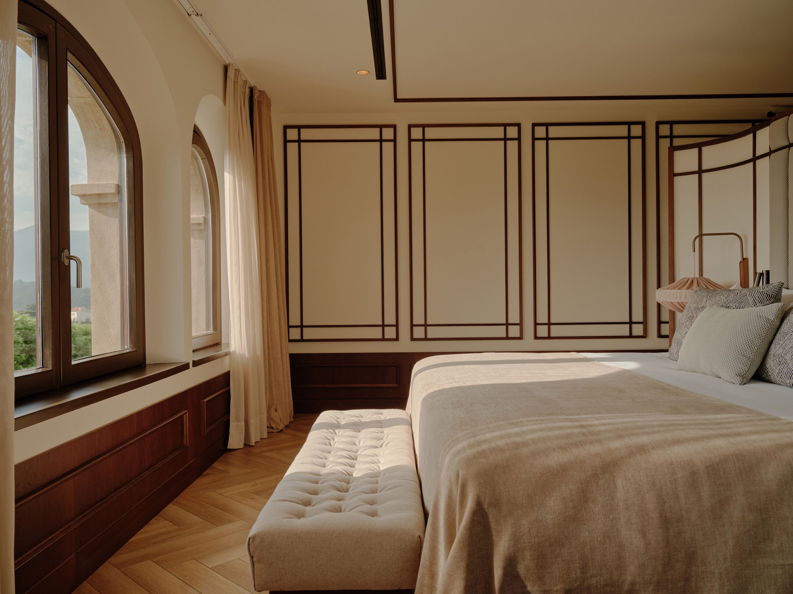 A bedroom with a large bed, a window with curtains, a wooden floor, and a view of the outdoors.