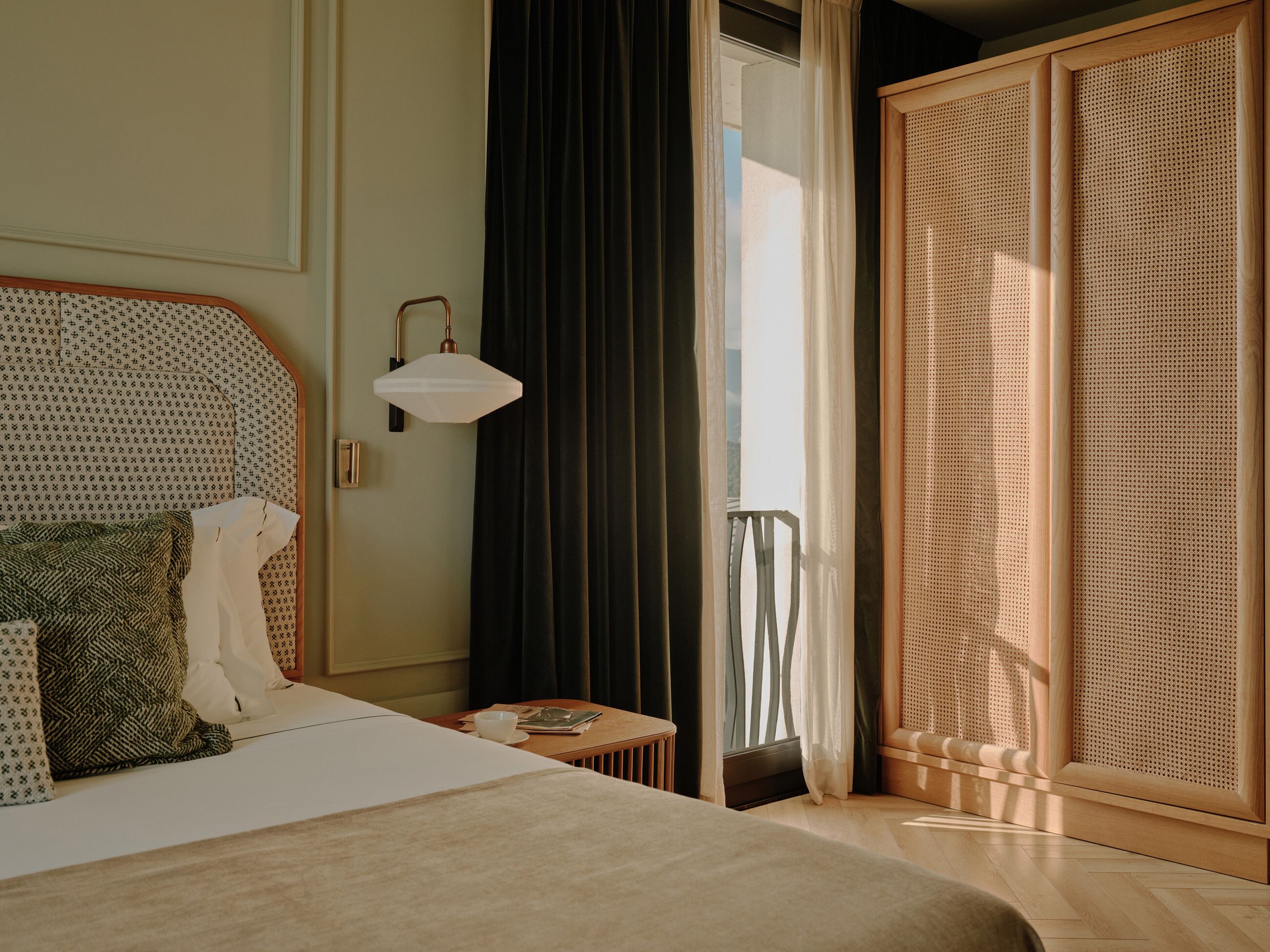 Een hotelkamer met een bed, een nachtkastje met een lamp, een raam met gordijnen en een houten kledingkast.