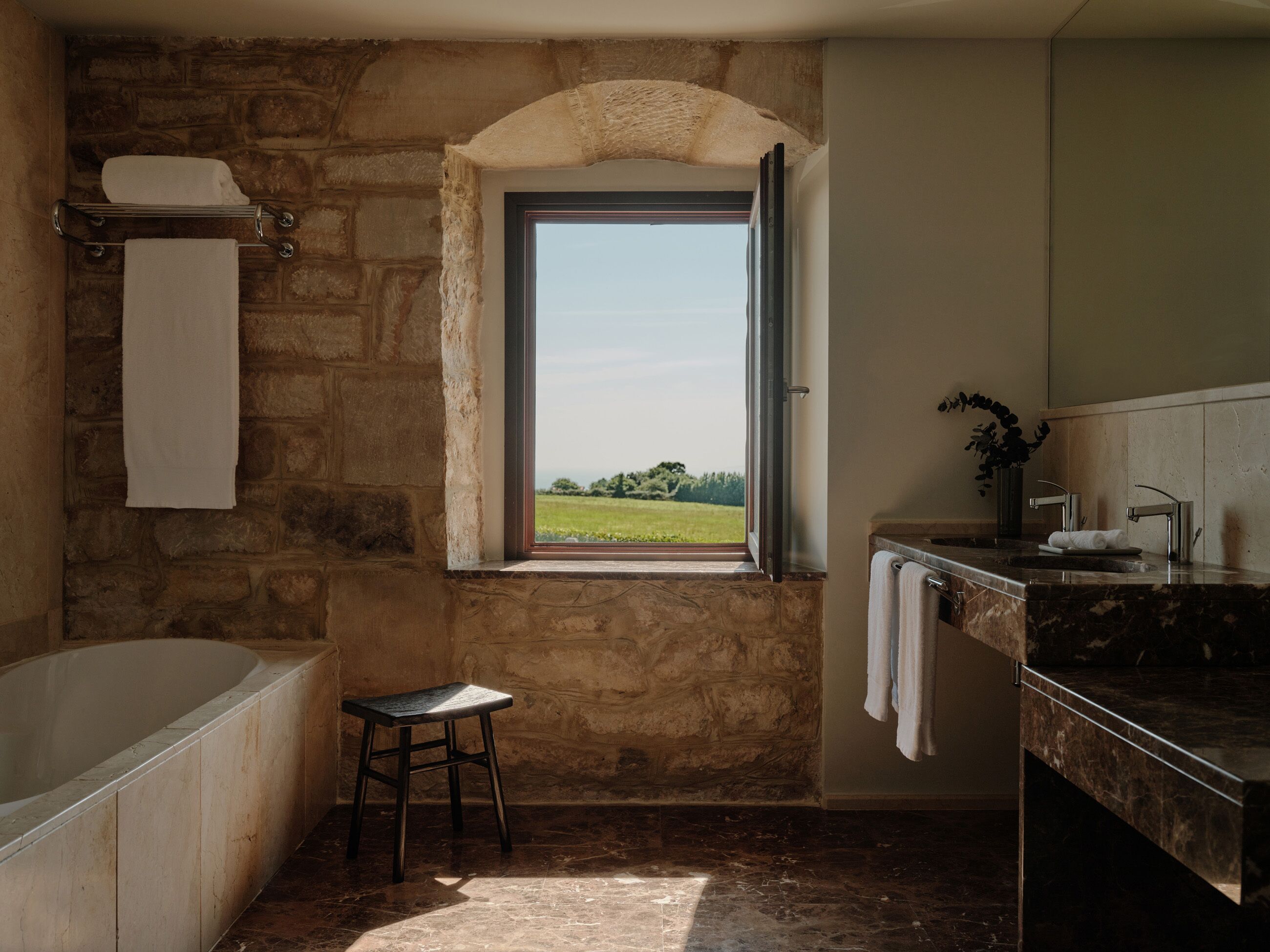 Un bany amb paret de pedra, una banyera, una finestra amb vista a un camp i un lavabo amb mirall.