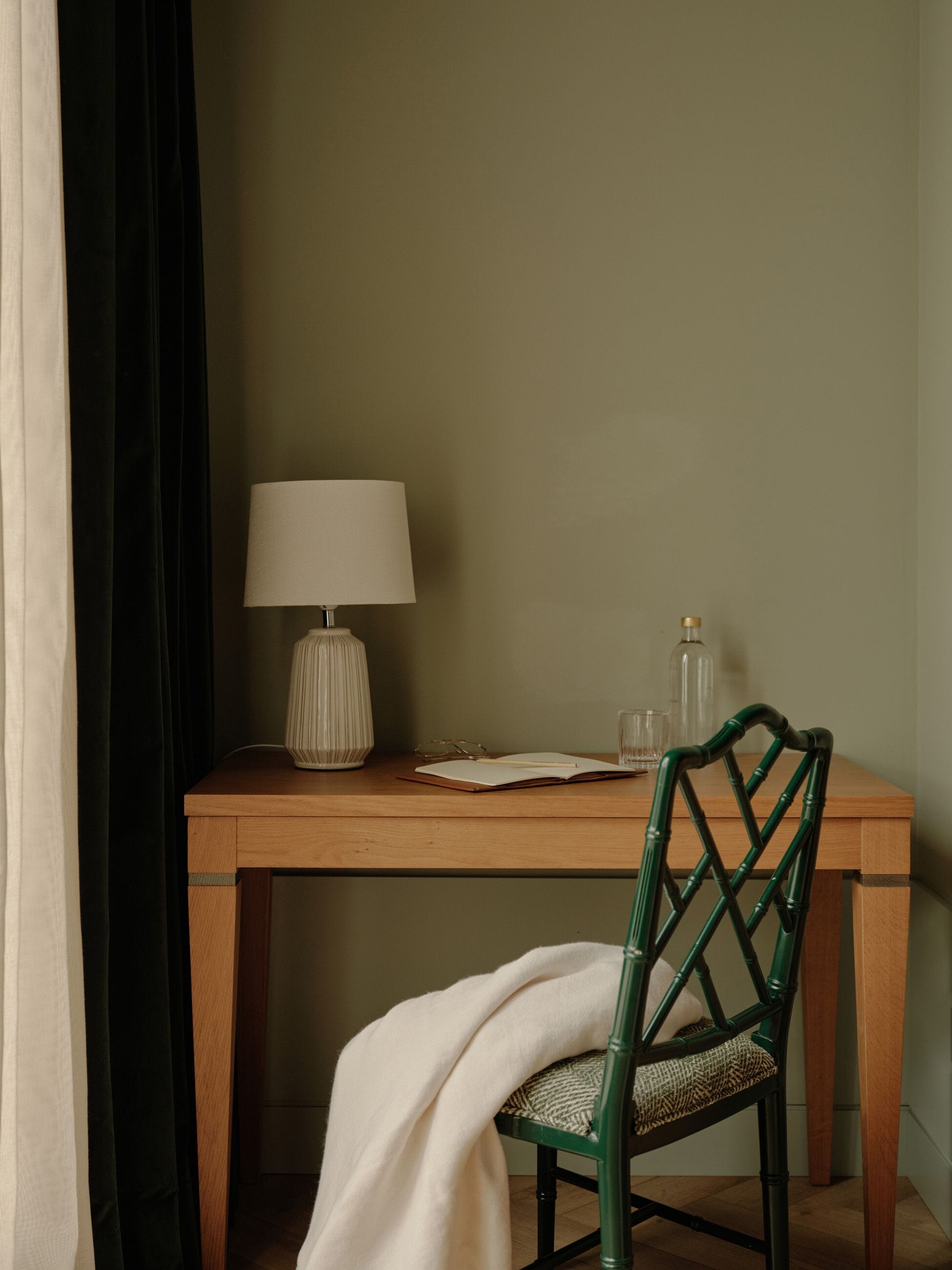 Une table en bois avec une lampe, des verres et une bouteille. On aperçoit partiellement une chaise recouverte d’un tissu blanc.