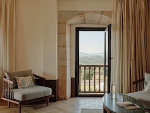 A room with a view of a mountainous landscape through a balcony door, a wooden chair with a cushion, a table with a glass and a book, and a window with curtains.