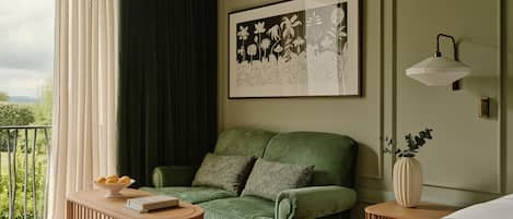 A room with a green sofa, a wooden side table, a framed floral artwork, a bedside lamp, and a view of greenery outside.