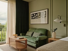 Una habitación con un sofá verde, una mesita de madera, un cuadro floral enmarco y una lámpara de noche, con vistas a la vegetación exterior.