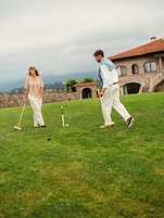 Dos personas jugando croquet en un campo de césped, con un muro de piedra y un edificio al fondo.
