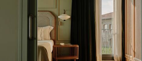 A hotel room with a bed, bedside table, lamp, and a view of the outside.