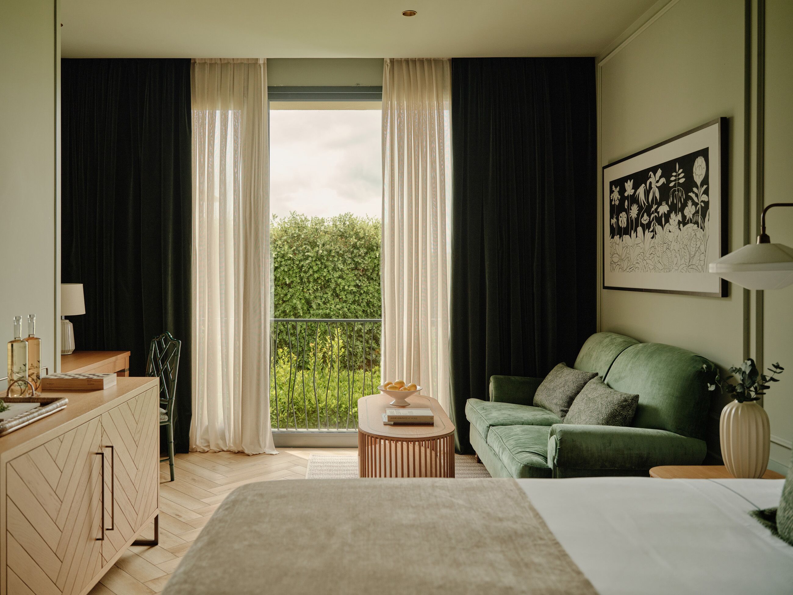 Un dormitorio con cama, sofá verde, cómoda de madera, mesita redonda y vistas a zonas verdes por la ventana.