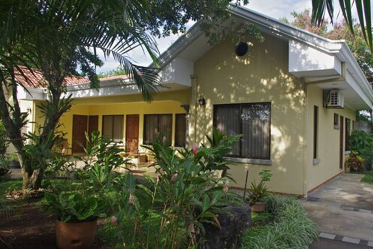 A two-story building with a yellow facade, a covered patio area, and a small garden with various plants.