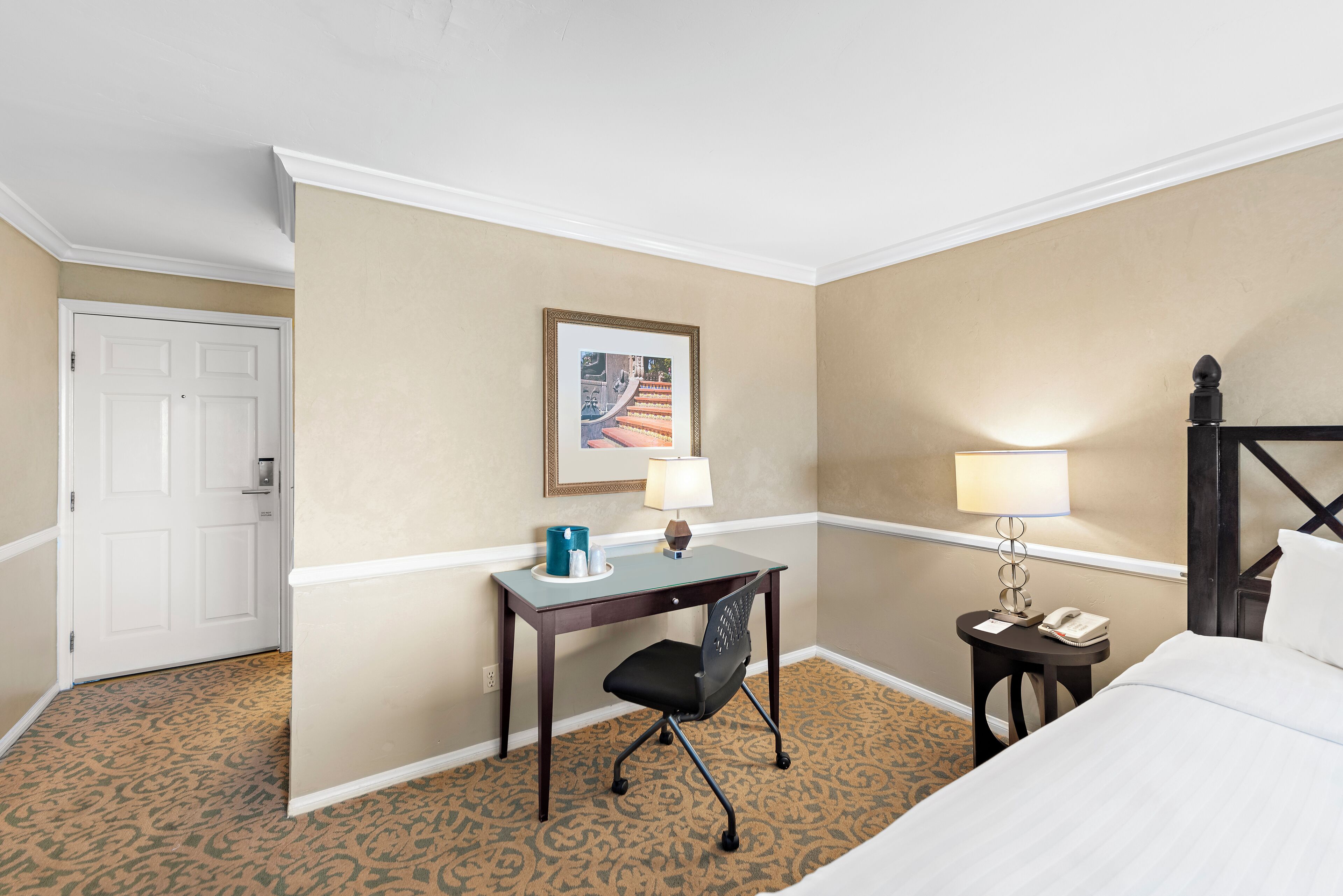 A hotel room with a bed, a desk with a chair, a small table with a lamp, a framed picture on the wall, and a door.