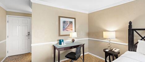 A hotel room with a bed, a desk with a chair, a small table with a lamp, a framed picture on the wall, and a door.