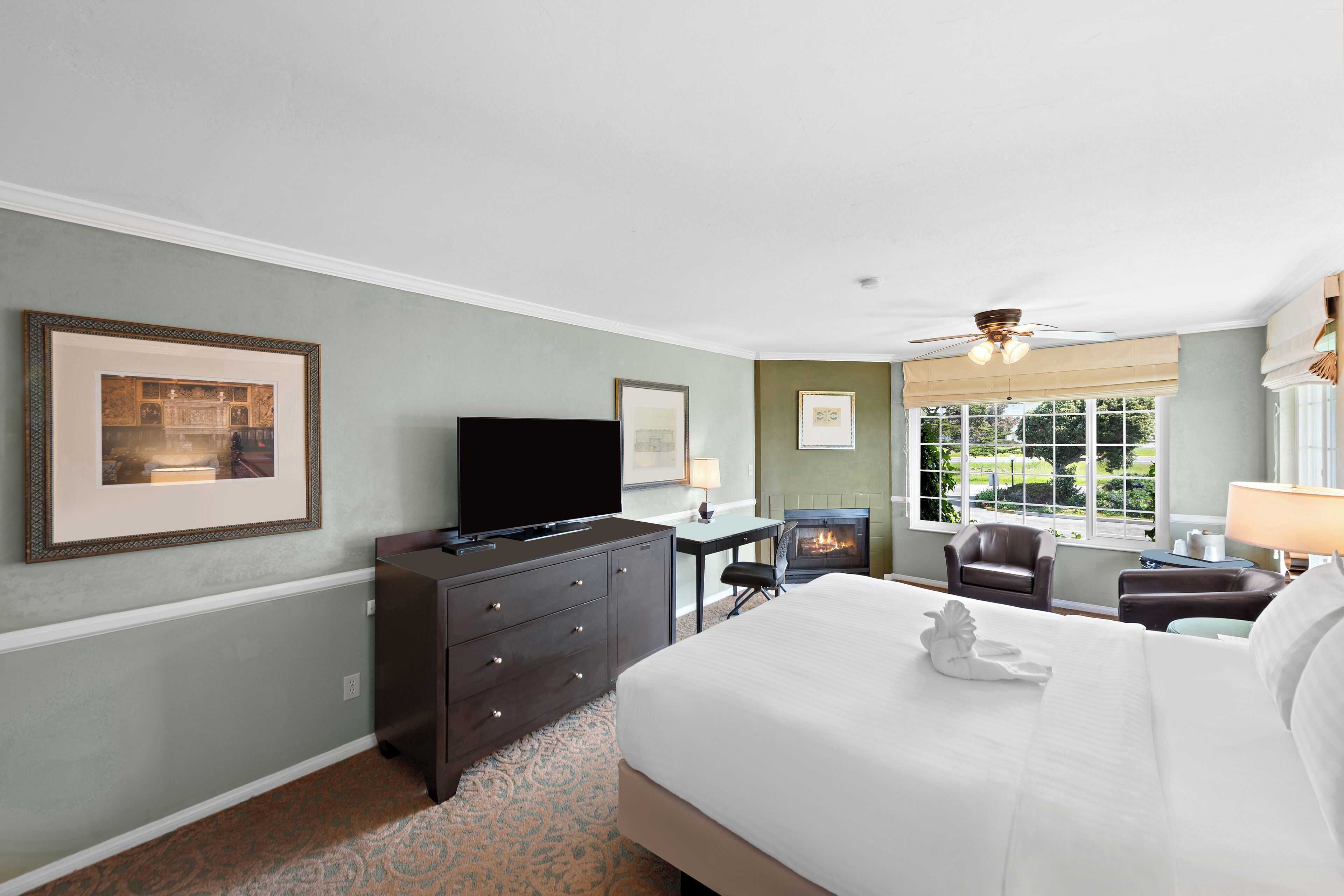 Ein Hotelzimmer mit einem Bett, einem Schminktisch, einem Fernseher, einem Schreibtisch, einem Sessel, einem Kamin und einem Fenster mit Blick auf Grünflächen.