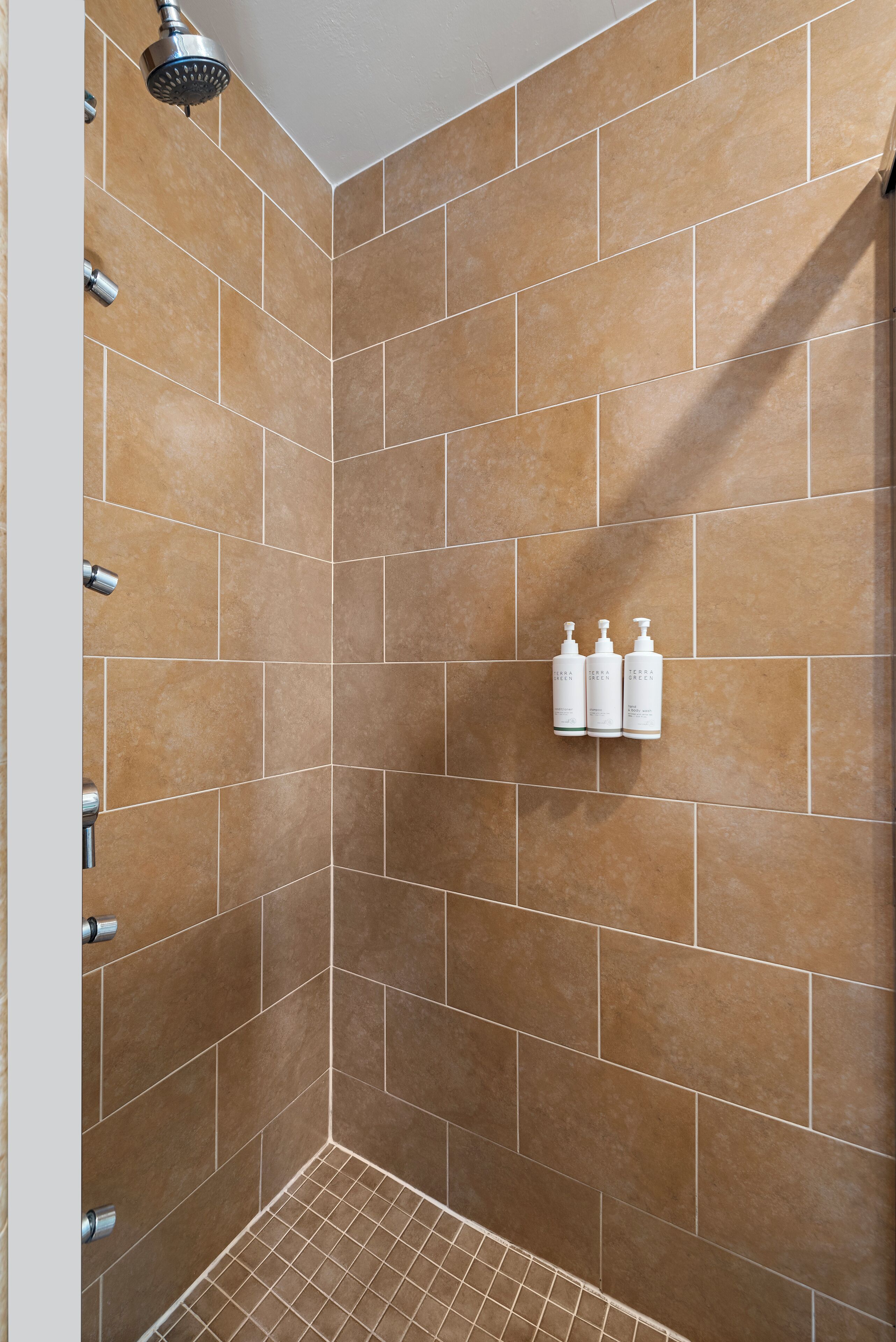Un área de ducha con azulejos marrones, cabezal de ducha y tres botellas de champú y acondicionador.