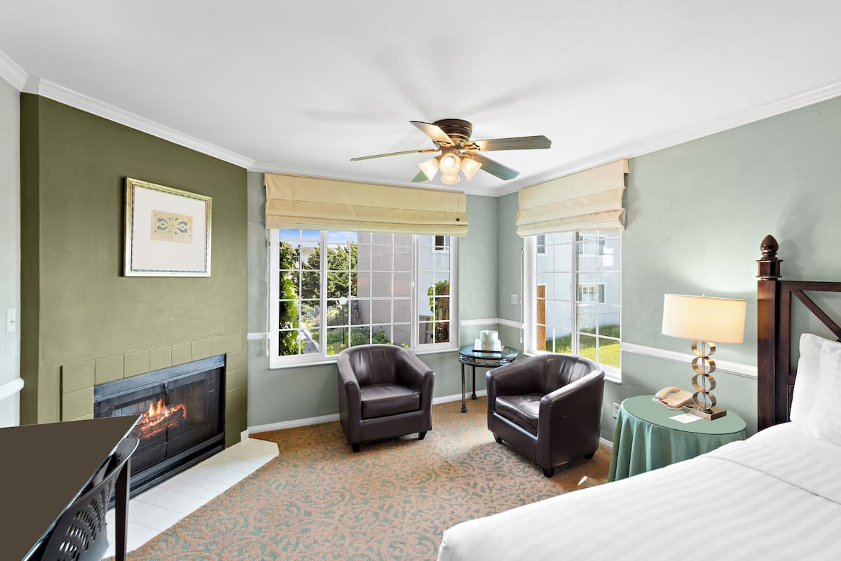 A hotel room with a fireplace, two leather armchairs, a bed, a round table with a lamp, and a ceiling fan.