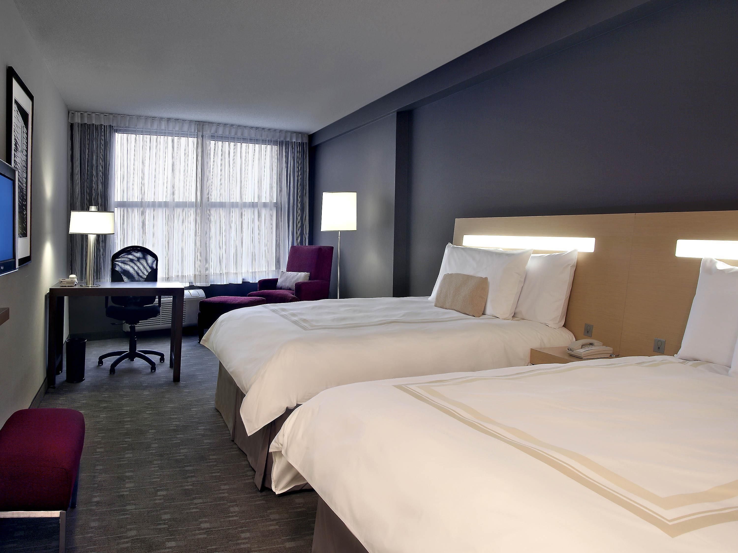A hotel room with two beds, a desk, a chair, and a large window with curtains.