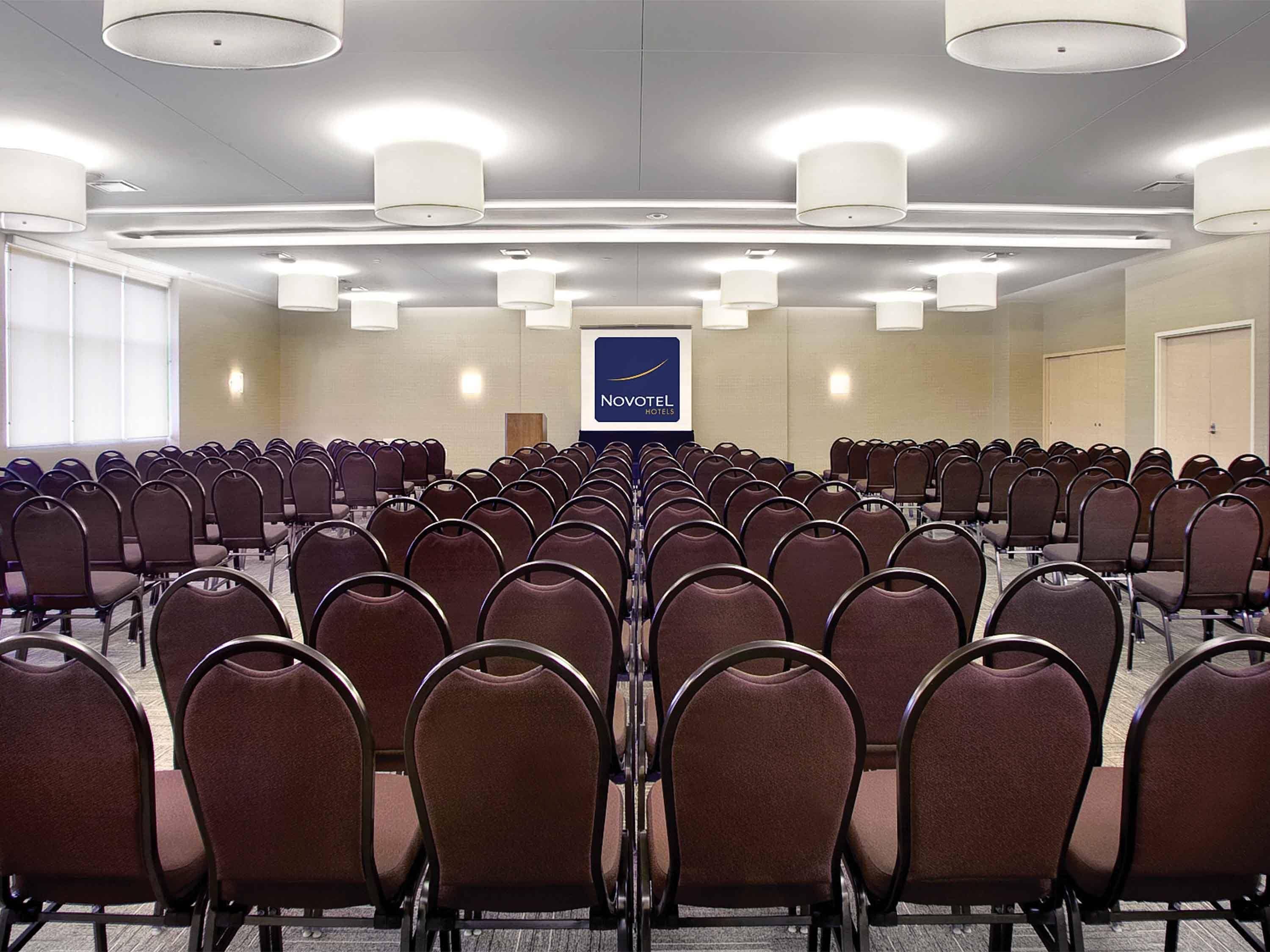 A conference room with rows of chairs, a podium, and a sign for Novotel Hotels.
