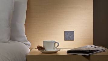 Una cama bien hecha con ropa blanca, una mesita de noche con una taza blanca, una galleta y un periódico doblado.