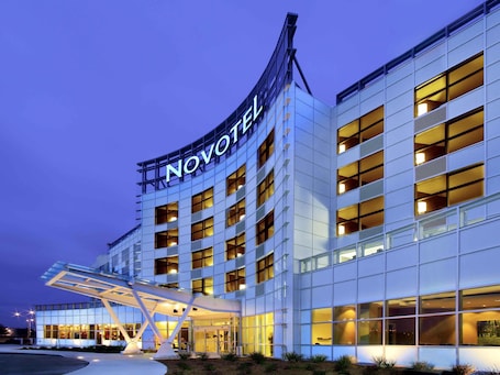 A modern hotel building with a prominent 'NOVOTEL' sign, multiple floors, and a covered entrance area.. Novotel Montreal Aeroport