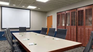 Un salón de conferencias con una larga mesa, seis sillas, una pantalla de proyector y una puerta con contraventanas.