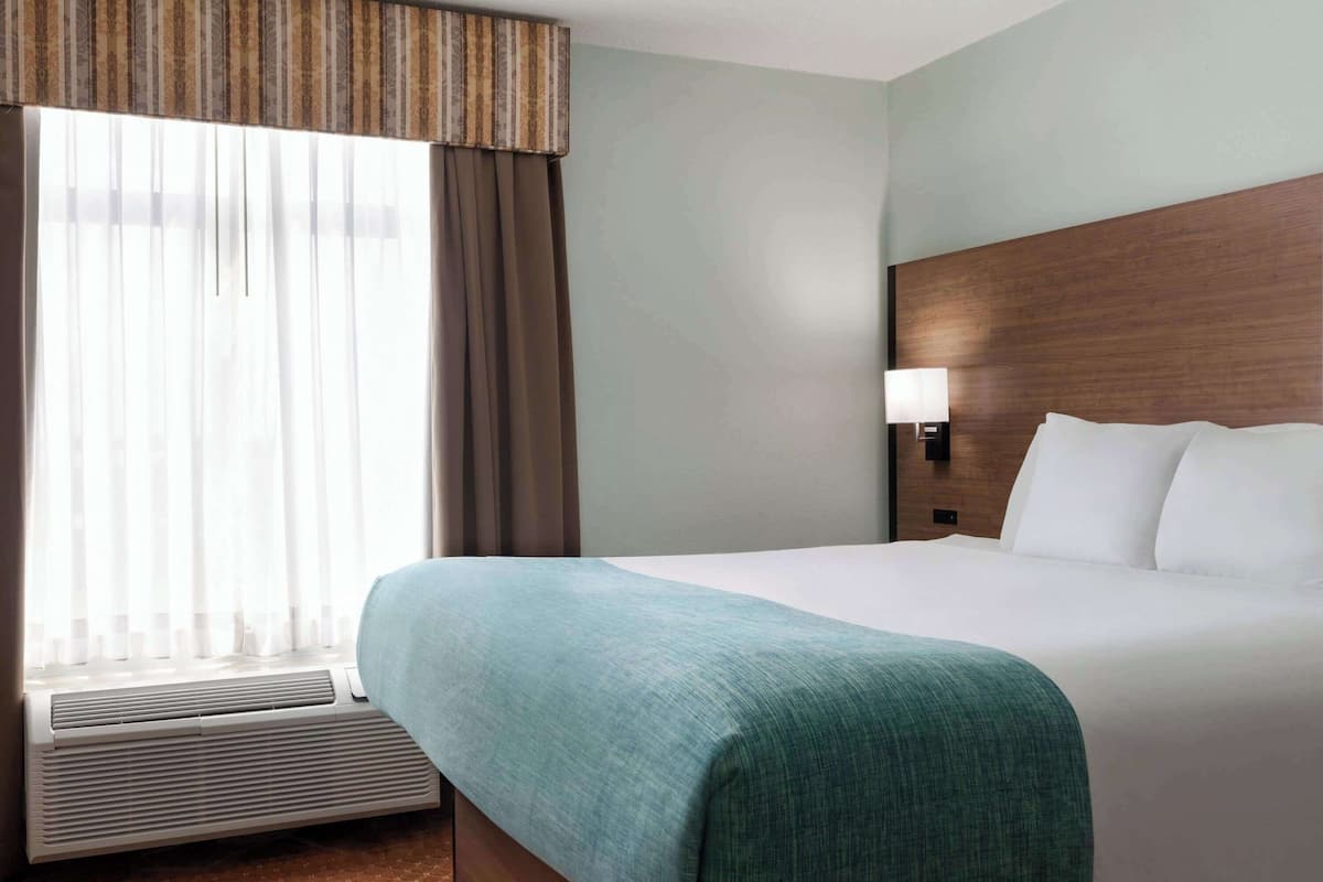 Une chambre d’hôtel avec un grand lit, une fenêtre avec des rideaux, une lampe fixée au mur et une tête de lit en bois.