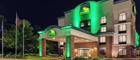 A Wingate by Wyndham hotel with a green-lit entrance, handicap parking, and a flag in the foreground.
