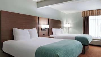 A hotel room with two beds, a wooden headboard, a nightstand with a lamp, and a large window with curtains.